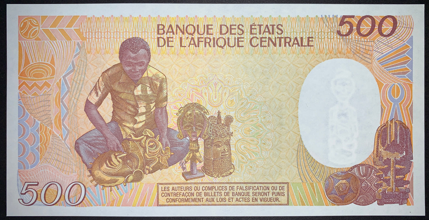 Reverse of the Central African Republic 500 Francs 1991 P-14d UNC banknote featuring a young artisan carving a traditional African tribal mask, surrounded by finished masks, sculptures, shields, and decorative cultural motifs. Authentic uncirculated collectible banknote.