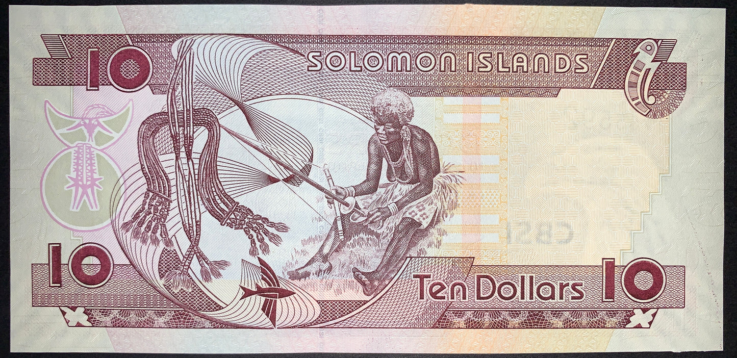Reverse view of a plum-purple Solomon Islands 10-dollar banknote in uncirculated condition, focusing on cultural heritage. The central vignette illustrates a traditional seated woman using a hand pump-drill to shape and manufacture historical shell money (shell bead currency). On the left side, a large string of completed shell money with woven tasselled ends hangs prominently against fine line geometric swirls. The denomination "Ten Dollars" is printed along the bottom right, with large numeral 10s framing