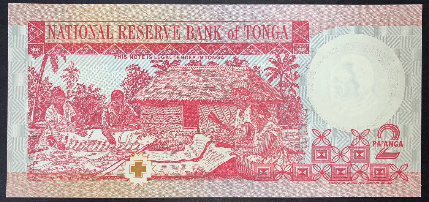 Reverse view of a red and light-pink Tonga 2 Pa'anga banknote in uncirculated condition. The main central vignette depicts an authentic outdoor cultural scene of four Tongan women sitting on the ground together, meticulously handcrafting and unrolling a long, traditional piece of Tapa cloth (Ngatu). A traditional thatched-roof stilt house and tropical palm trees form the background. The text "NATIONAL RESERVE BANK of TONGA" is printed in a red banner along the top border.