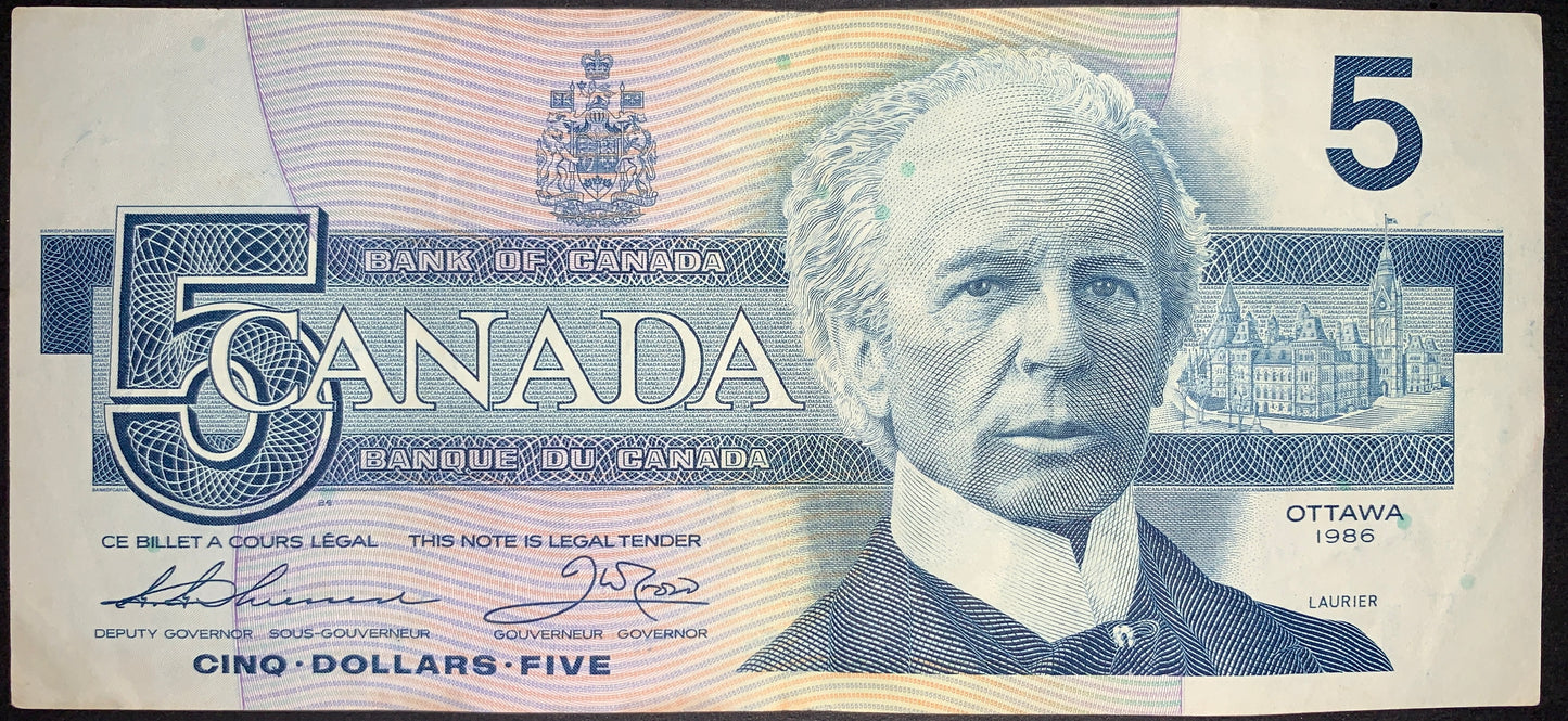 Obverse view of a crisp 1986 Canadian 5-dollar banknote from the Birds of Canada series featuring a portrait of Sir Wilfrid Laurier. The bill showcases detailed blue and multi-color security patterns, the official Coat of Arms of Canada, and an illustration of the Parliament buildings in Ottawa. The signatures of the Deputy Governor and Governor are clearly visible at the bottom left, alongside a prominent large numeral 5 on the right.