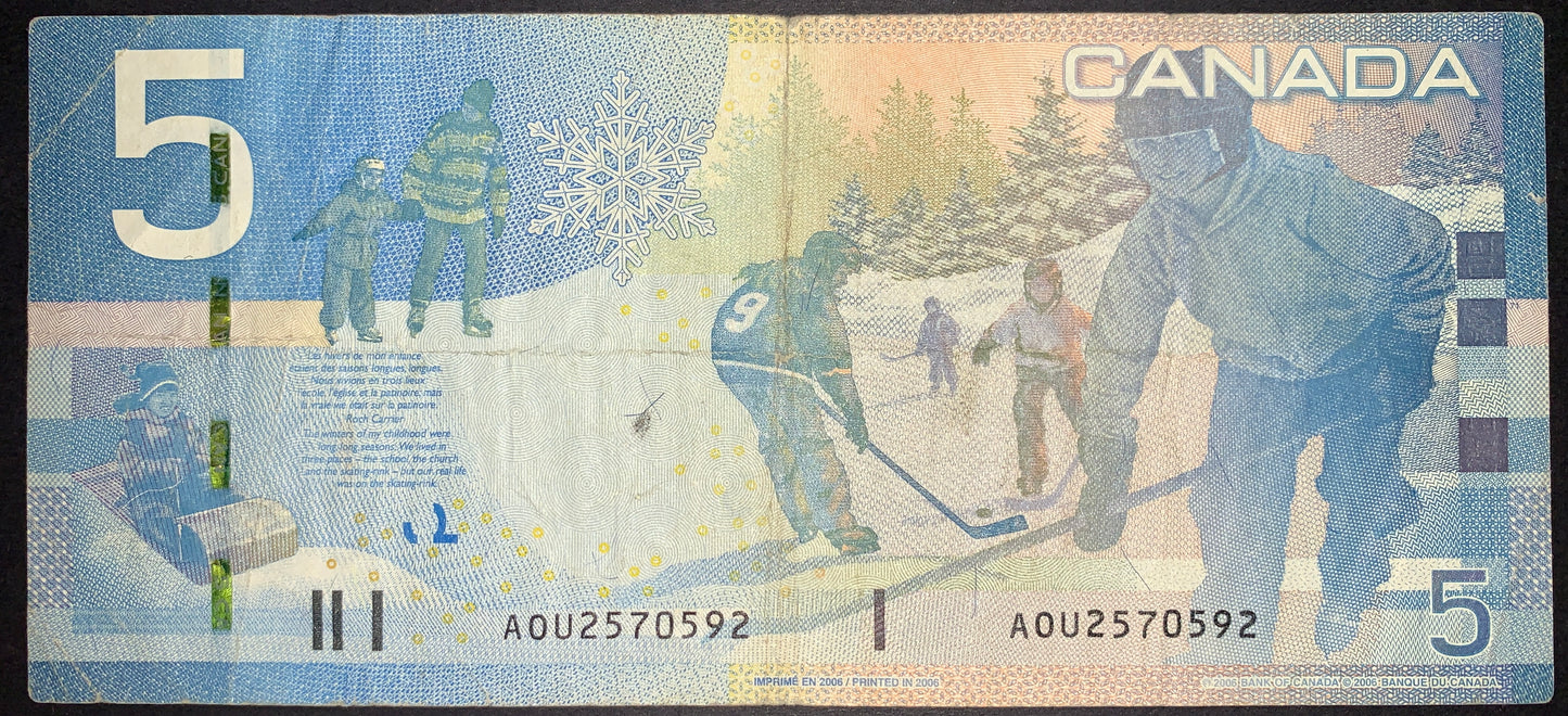 Reverse view of a 2006 Canadian 5-dollar banknote from the Canadian Journey series highlighting the Children at Play theme. The design features detailed illustrations of children playing ice hockey, outdoor ice skating, and tobogganing in a winter landscape, alongside a large stylized snowflake. A bilingual excerpt from Roch Carrier's short story is printed on the left. The serial number AOU2570592 is printed twice along the bottom border in black ink, with large numeral 5s in the top-left and bottom-right 