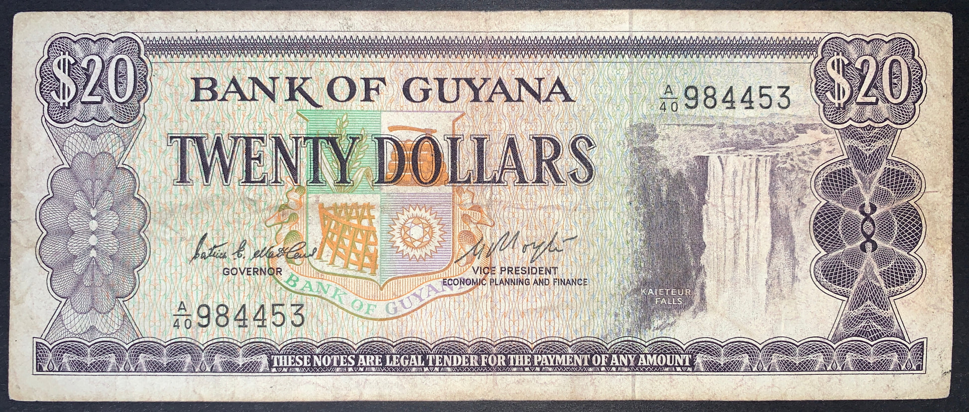 Obverse view of a purple and multi-colored Guyana 20-dollar banknote featuring the majestic Kaieteur Falls on the right side. The text "BANK OF GUYANA" and "TWENTY DOLLARS" is prominently displayed in the center above a large, colorful official Coat of Arms. The serial number A/40 984453 is printed in black ink on the lower-left and upper-right. Intricate purple geometric guilloche patterns and rosettes frame the note, with large "$20" numerals in the top corners.