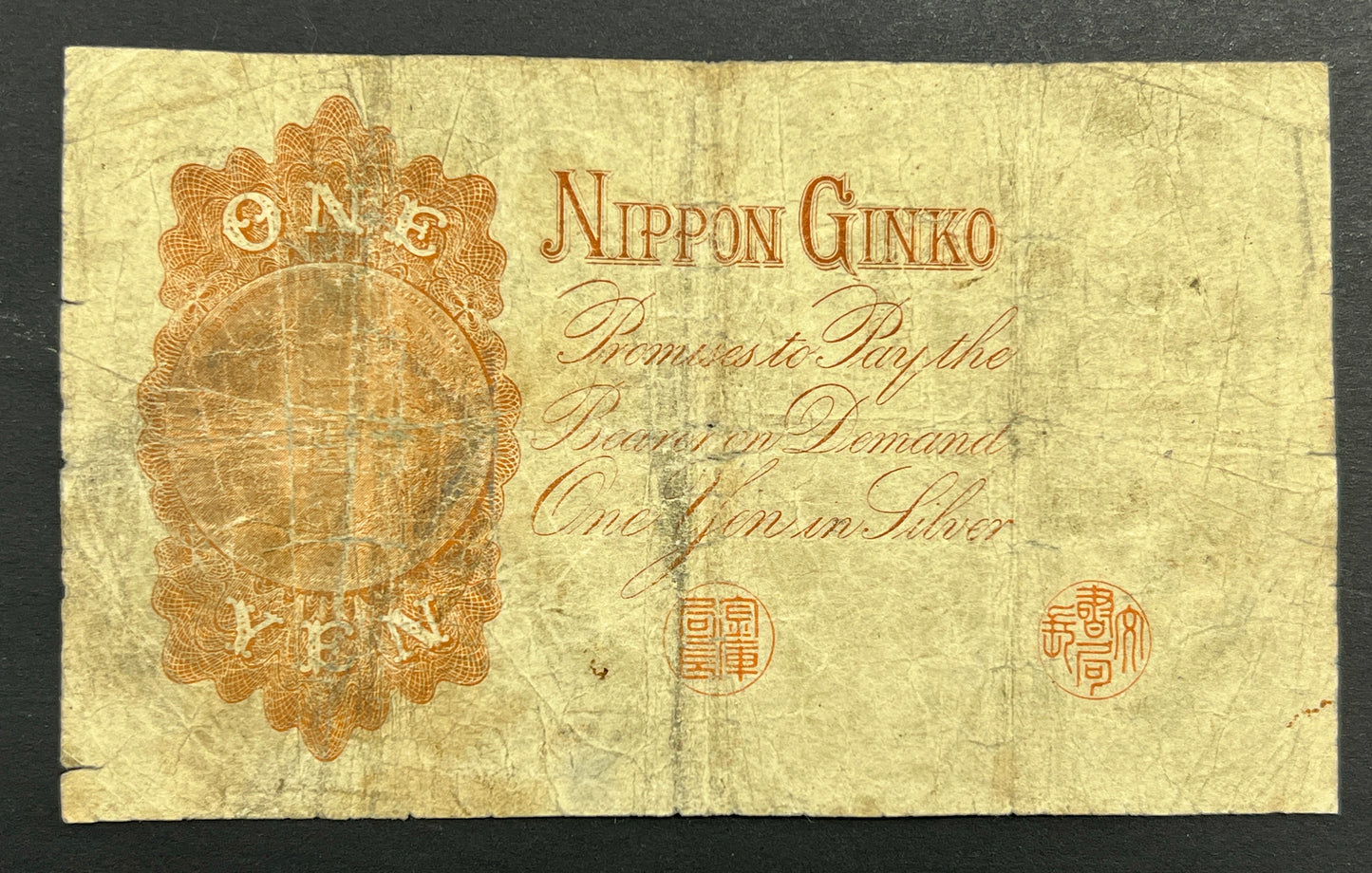 Back view of a heavily circulated 1889 Japan 1 Yen banknote showcasing an ornate orange engraving that reads "One Yen in Silver" by Nippon Ginko.
