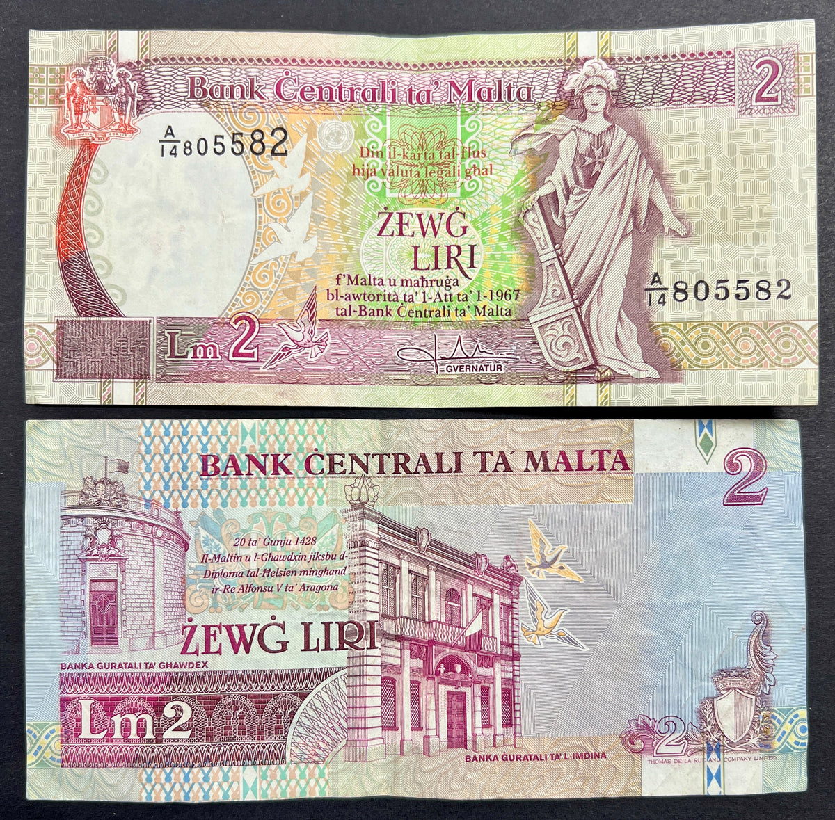 Front view of a circulated 1989 Malta 2 Liri banknote featuring the allegorical figure of Melita holding a ship's rudder and the Maltese coat of arms. Back view of a vintage 1989 Malta 2 Liri banknote showcasing detailed engravings of the historic Banca Giuratale buildings in Mdina and Gozo.