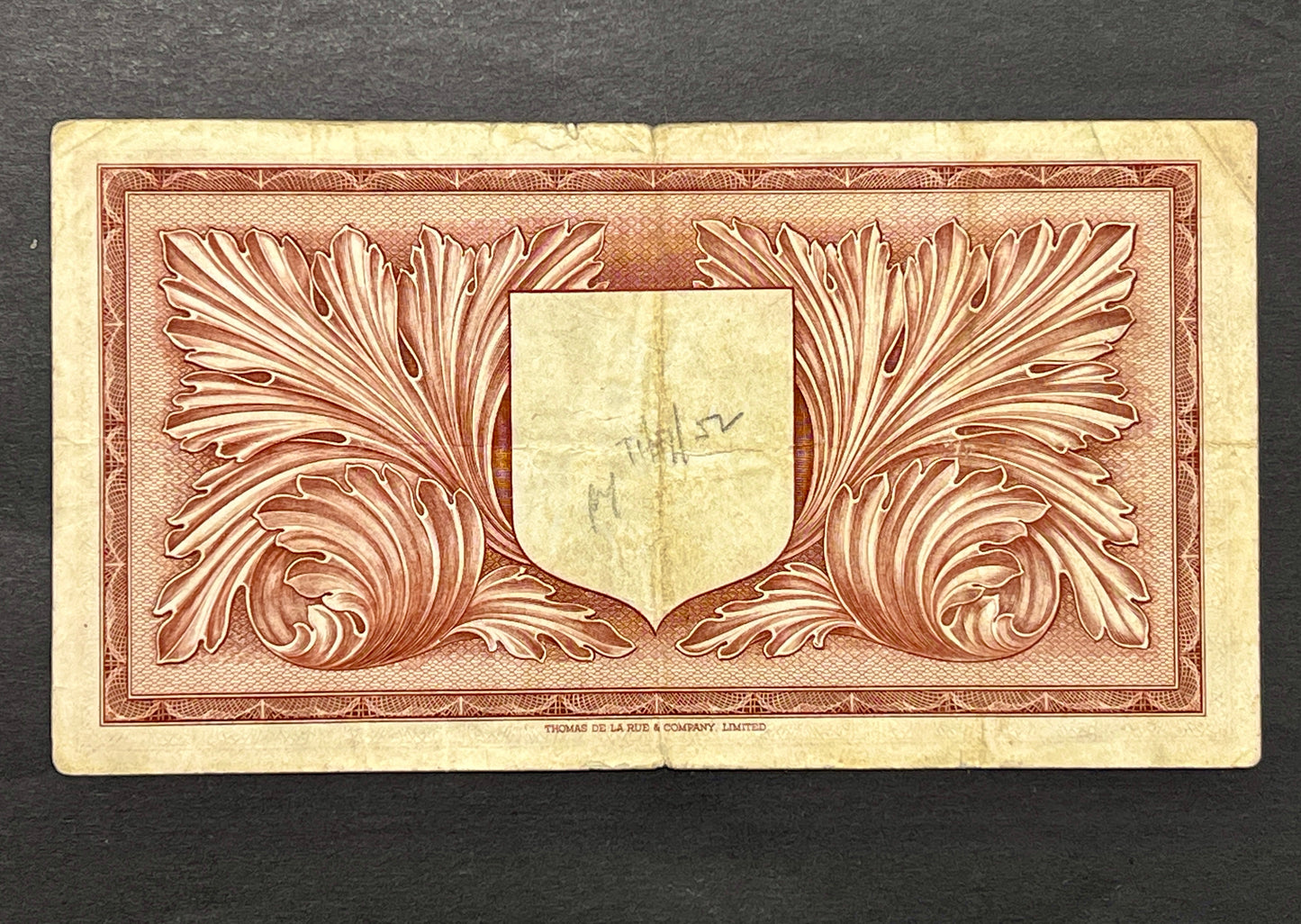 Back view of a 1949 Malta 1 Pound banknote showcasing intricate acanthus leaves and a central shield, printed by Thomas De La Rue.