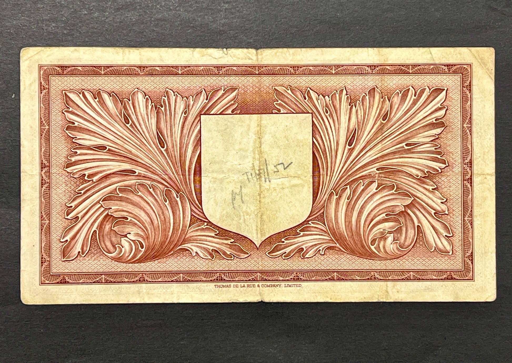 Back view of a 1949 Malta 1 Pound banknote showcasing intricate acanthus leaves and a central shield, printed by Thomas De La Rue.