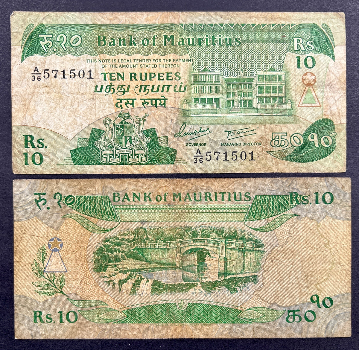 Front view of a circulated 1985 Mauritius 10 Rupees banknote featuring the national coat of arms and the historic Government House building in Port Louis. Back view of a vintage 1985 Mauritius 10 Rupees banknote showcasing a detailed engraving of a classic stone bridge spanning over a scenic river and waterfall.