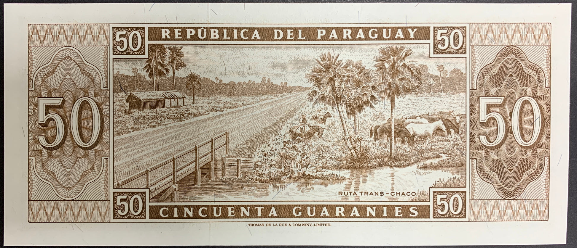 Reverse of a Paraguay 50 Guaraníes banknote (P-197b) featuring an engraved rural landscape of the Trans-Chaco Route in Paraguay. The scene includes a wooden bridge crossing a waterway, palm trees, open countryside, cattle grazing near the water, and a small structure in the distance. Large denomination numerals appear on both sides, surrounded by detailed ornamental patterns and fine-line engraving. Actual serial numbers may vary as multiple banknotes are available.