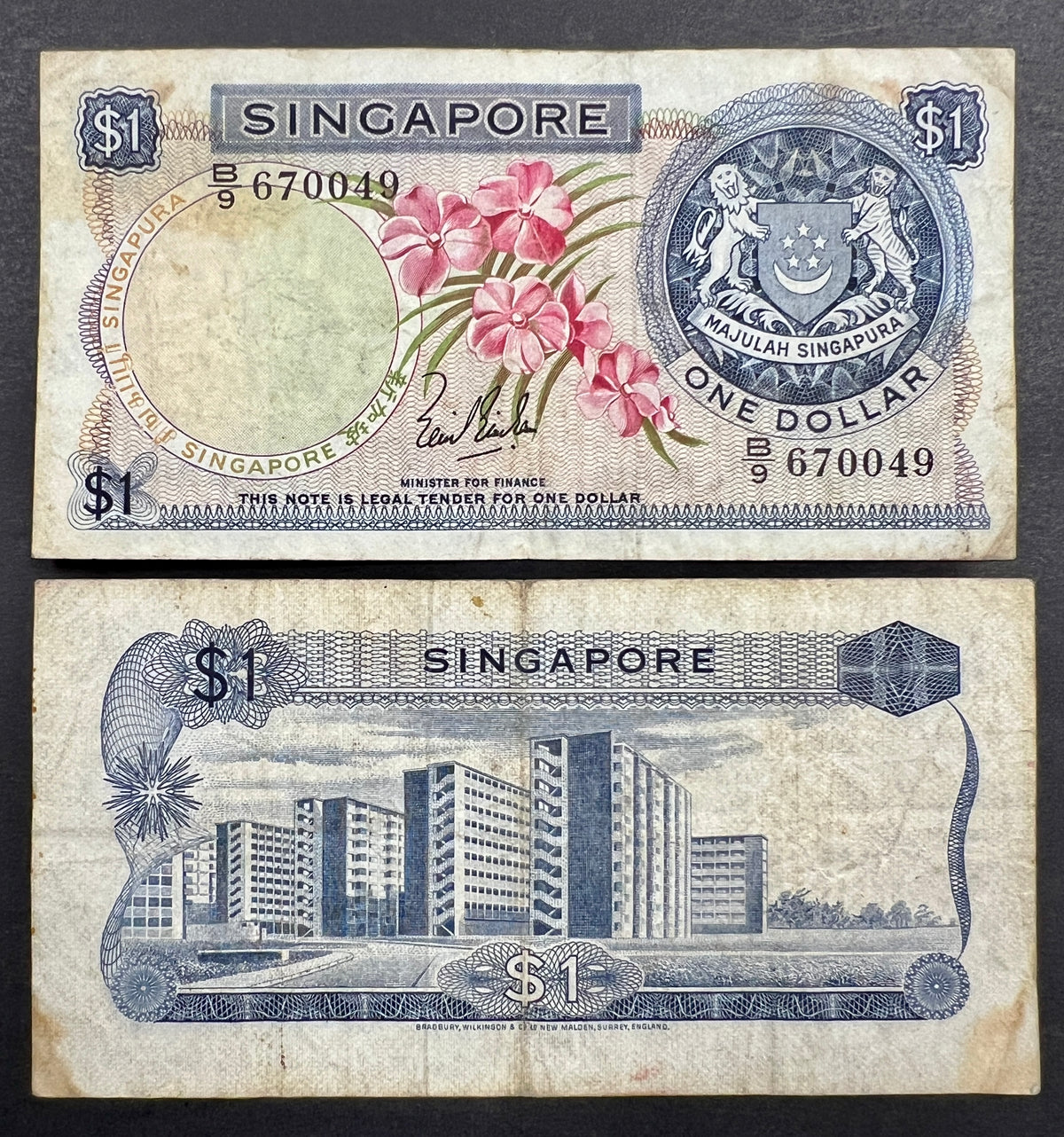 Front view of a circulated 1967 Singapore 1 Dollar banknote featuring a beautiful pink Vanda Joaquim orchid and the Singaporean national coat of arms.
Back view of a vintage 1967 Singapore 1 Dollar banknote showcasing a detailed engraving of modern high-rise public housing apartments.
