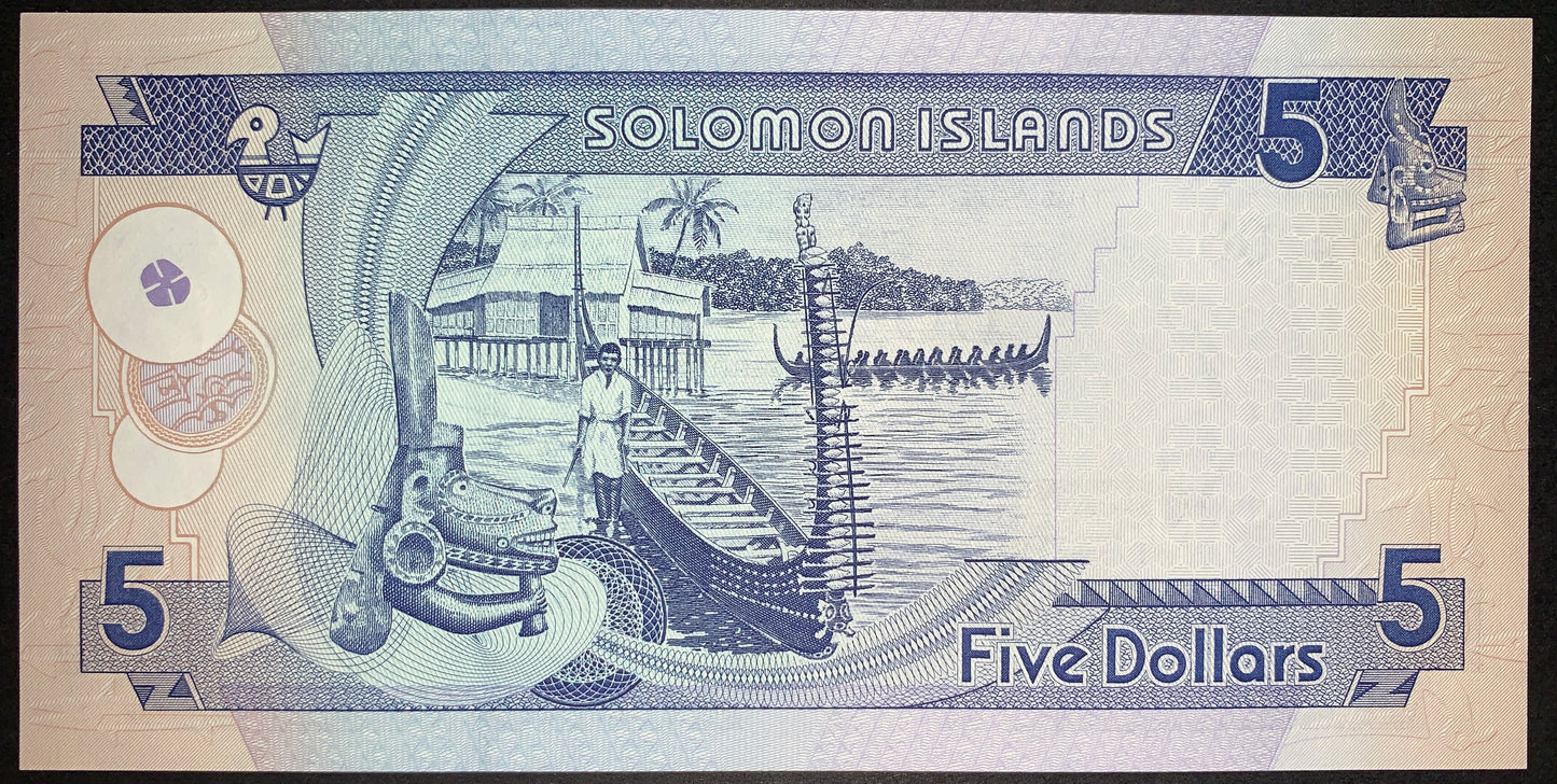 Reverse view of a Solomon Islands 5-dollar banknote in uncirculated condition, printed in deep blue ink against a light background. The central design beautifully illustrates a local coastal village scene featuring a man standing by a long traditional canoe on the shore, a village house on stilts over the water, and another large outrigger longboat with multiple paddlers out at sea. The left foreground showcases an intricate cultural wood carving ornament set against swirling guilloche line patterns, while 