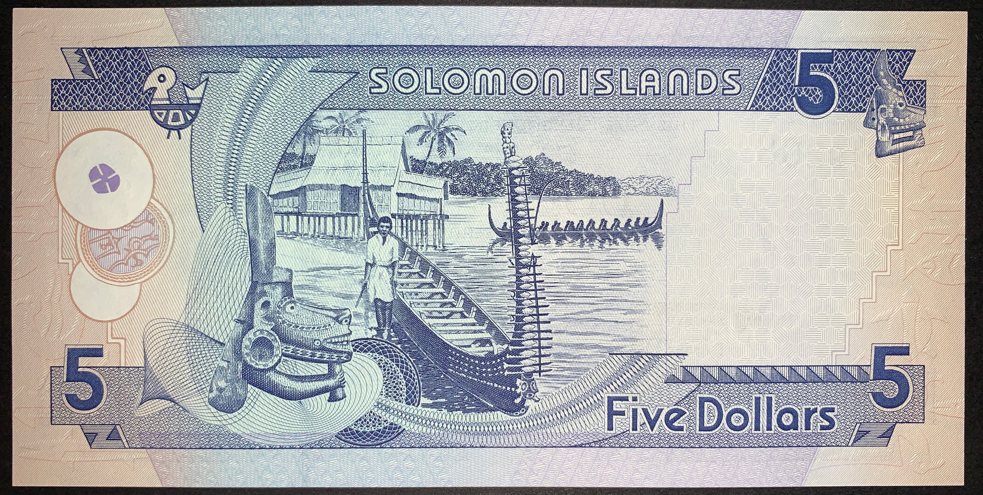 Reverse view of a Solomon Islands 5-dollar banknote in uncirculated condition, printed in deep blue ink against a light background. The central design beautifully illustrates a local coastal village scene featuring a man standing by a long traditional canoe on the shore, a village house on stilts over the water, and another large outrigger longboat with multiple paddlers out at sea. The left foreground showcases an intricate cultural wood carving ornament set against swirling guilloche line patterns, while 
