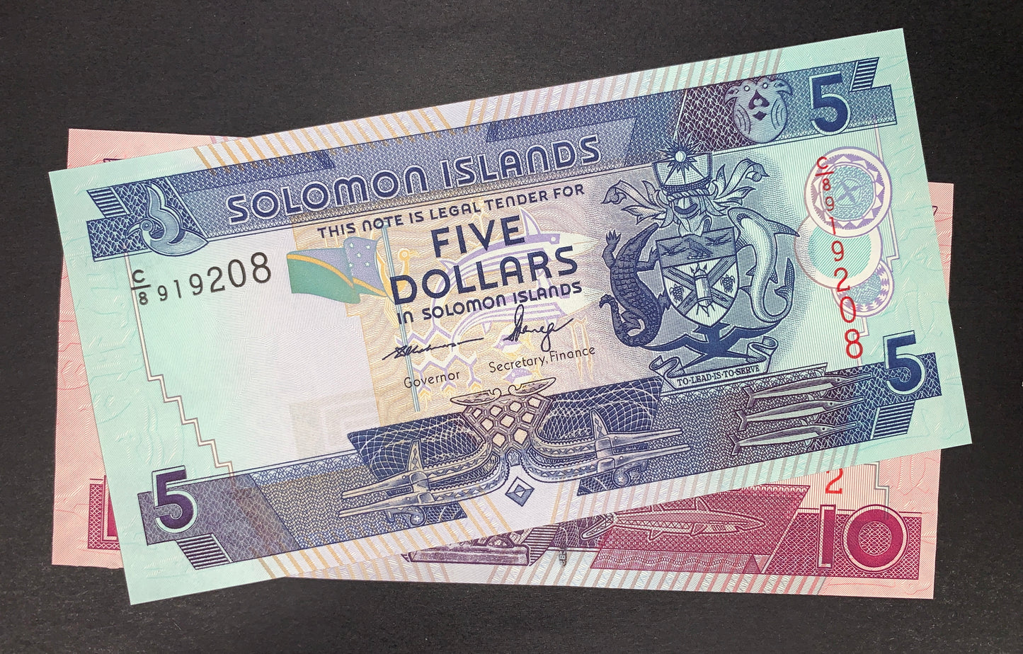 Obverse view of a Solomon Islands uncirculated banknote bundle showing the green and blue 5-dollar note stacked on top of a pink 10-dollar note. The 5-dollar note prominently features the national Coat of Arms on the right, a small flag in the center, and the signatures of Governor Rarawa and Secretary of Finance Fanega. The black serial number C/8 919208 is printed on the left, and a vertical red serial number is on the right. Large numeral 5s are positioned in multiple corners against intricate geometric 