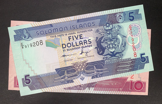 Obverse view of a Solomon Islands uncirculated banknote bundle showing the green and blue 5-dollar note stacked on top of a pink 10-dollar note. The 5-dollar note prominently features the national Coat of Arms on the right, a small flag in the center, and the signatures of Governor Rarawa and Secretary of Finance Fanega. The black serial number C/8 919208 is printed on the left, and a vertical red serial number is on the right. Large numeral 5s are positioned in multiple corners against intricate geometric 