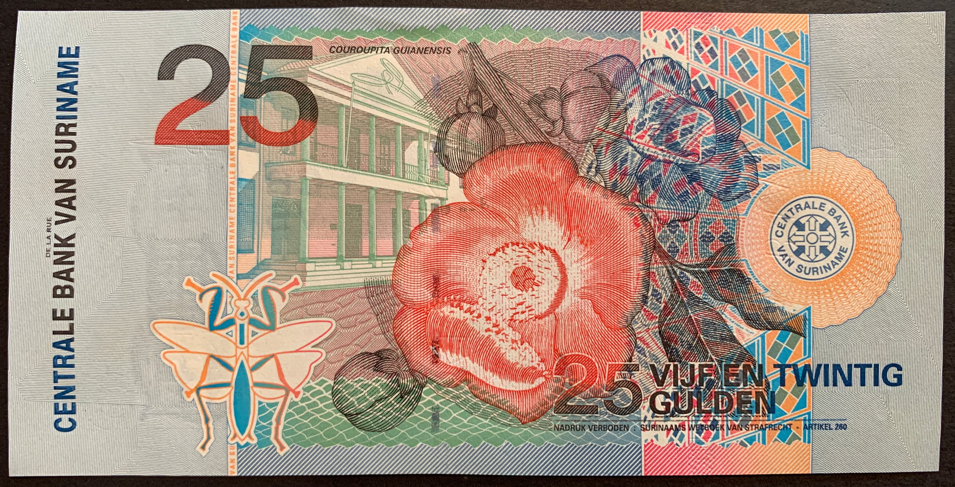 back of Suriname 25 Gulden 2000 P-148 collectible banknote with Cannonball flower (Couroupita guianensis) over Central Bank Building, stylized mantis
