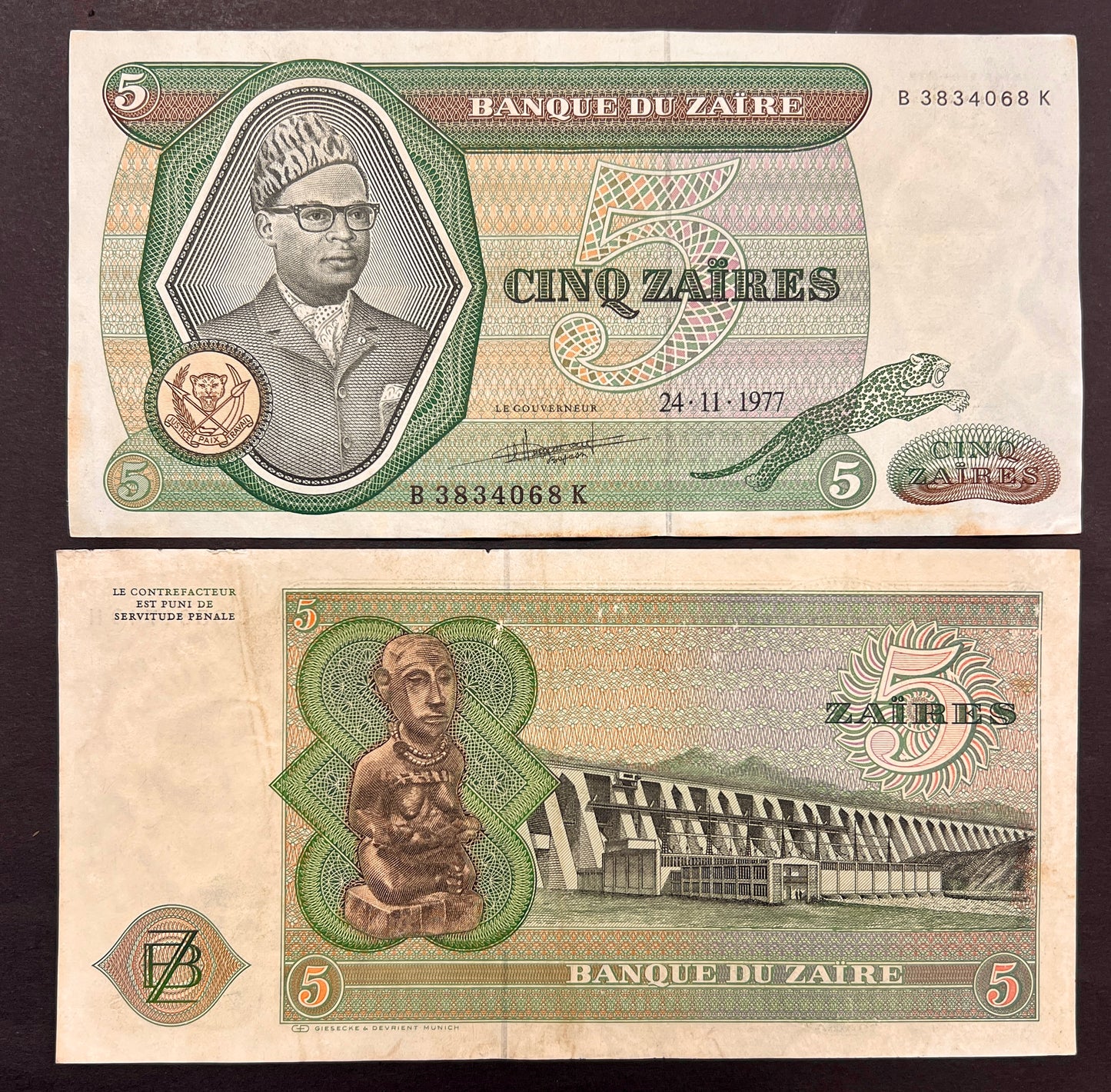 Front view of a circulated 1977 Zaire 5 Zaires banknote featuring a portrait of President Mobutu Sese Seko wearing his signature leopard-print hat, alongside a leaping leopard. Back view of a vintage 1977 Zaire 5 Zaires banknote showcasing a traditional carved wooden statue and an engraving of the massive Inga Dam.
