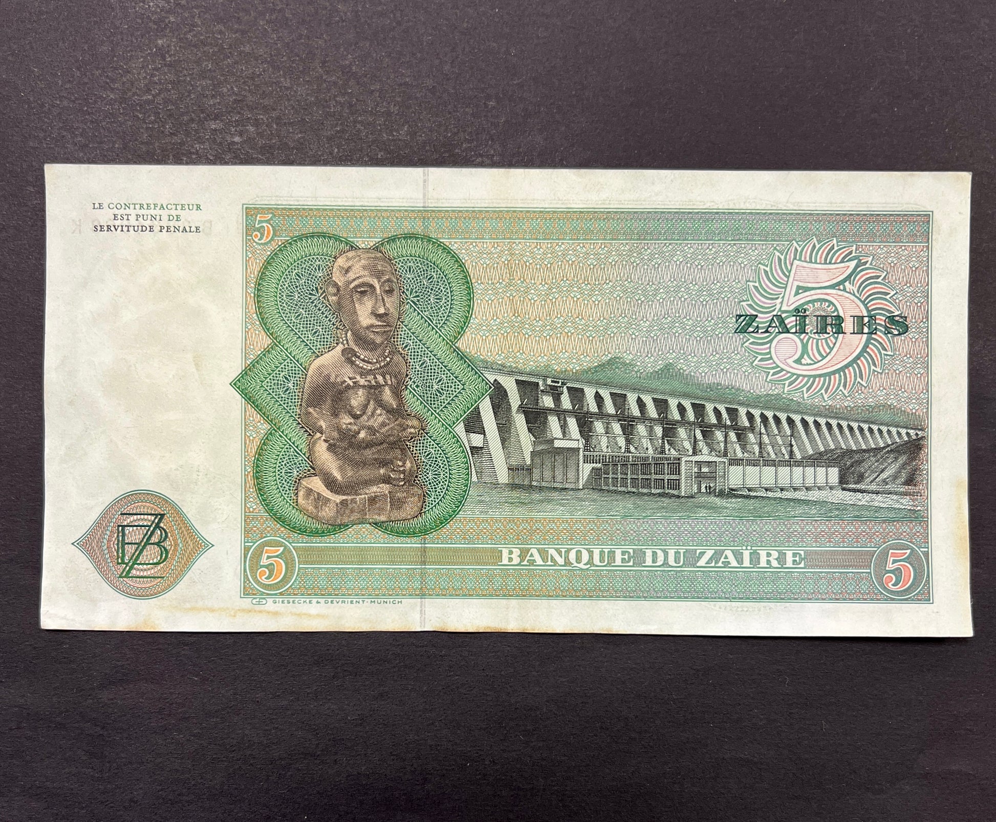 Back view of a vintage 1977 Zaire 5 Zaires banknote showcasing a traditional carved wooden statue and an engraving of the massive Inga Dam.
