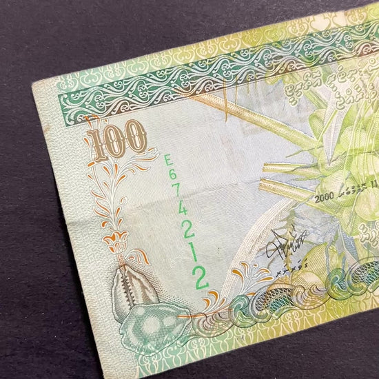 Front view of a circulated 2000 Maldives 100 Rufiyaa banknote featuring a traditional Dhoni sailing boat, a coconut palm, and a cowrie shell.
